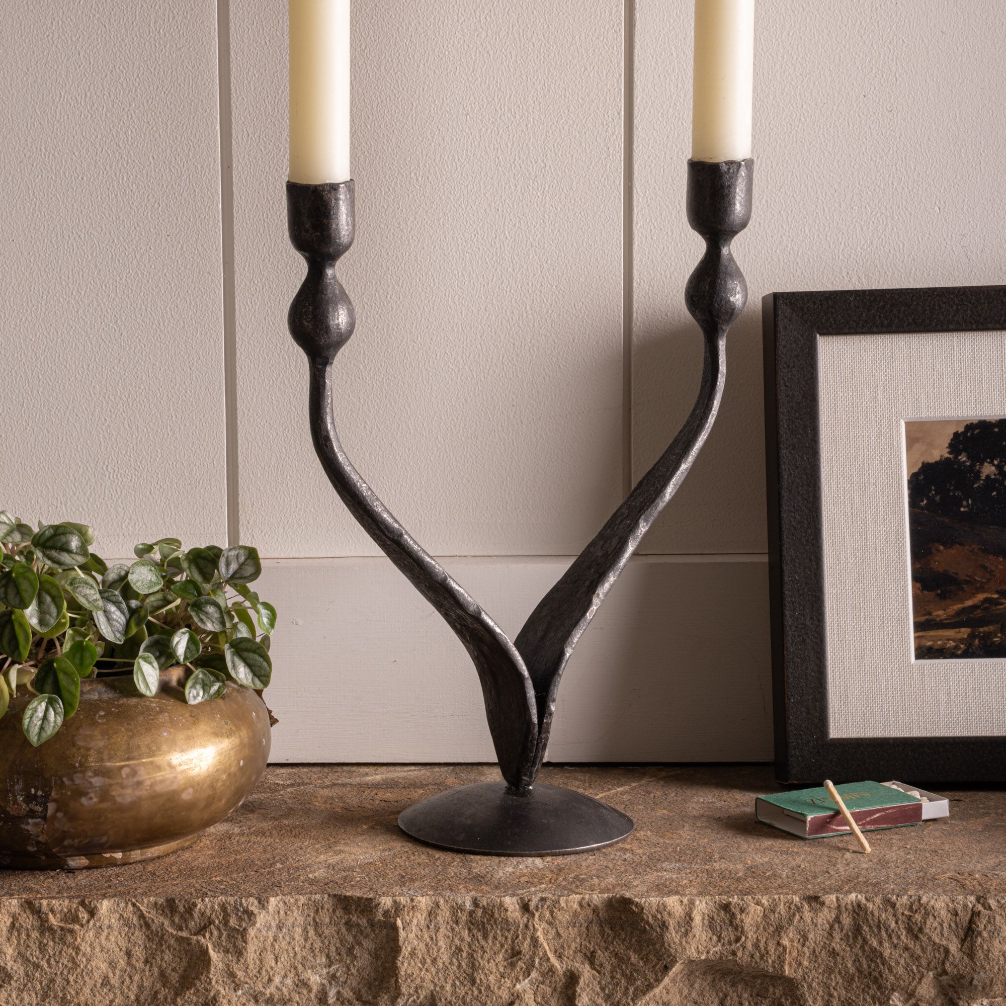 Black wrought iron candelabra with two cream taper candles on a stone mantel beside a brass planter and framed art.