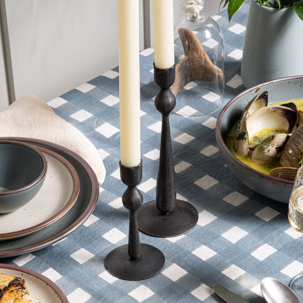 Two forged candle holders with cream tapers on a table with a blue-grey tablecloth with blue-grey and cream ceramic dinnerware