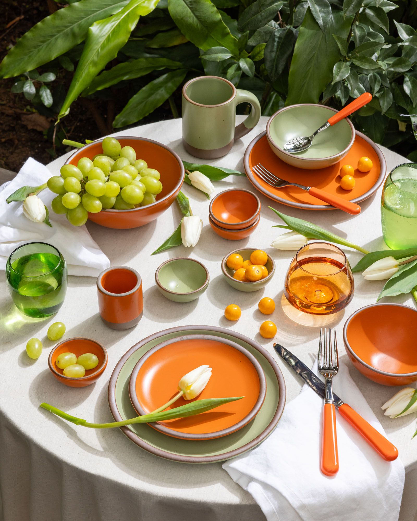 In an outdoor setting, a table is covered in ceramic dinnerware in a bold orange and calming sage green colors, surrounded by glasses, flatware and flowers in the same color palette.