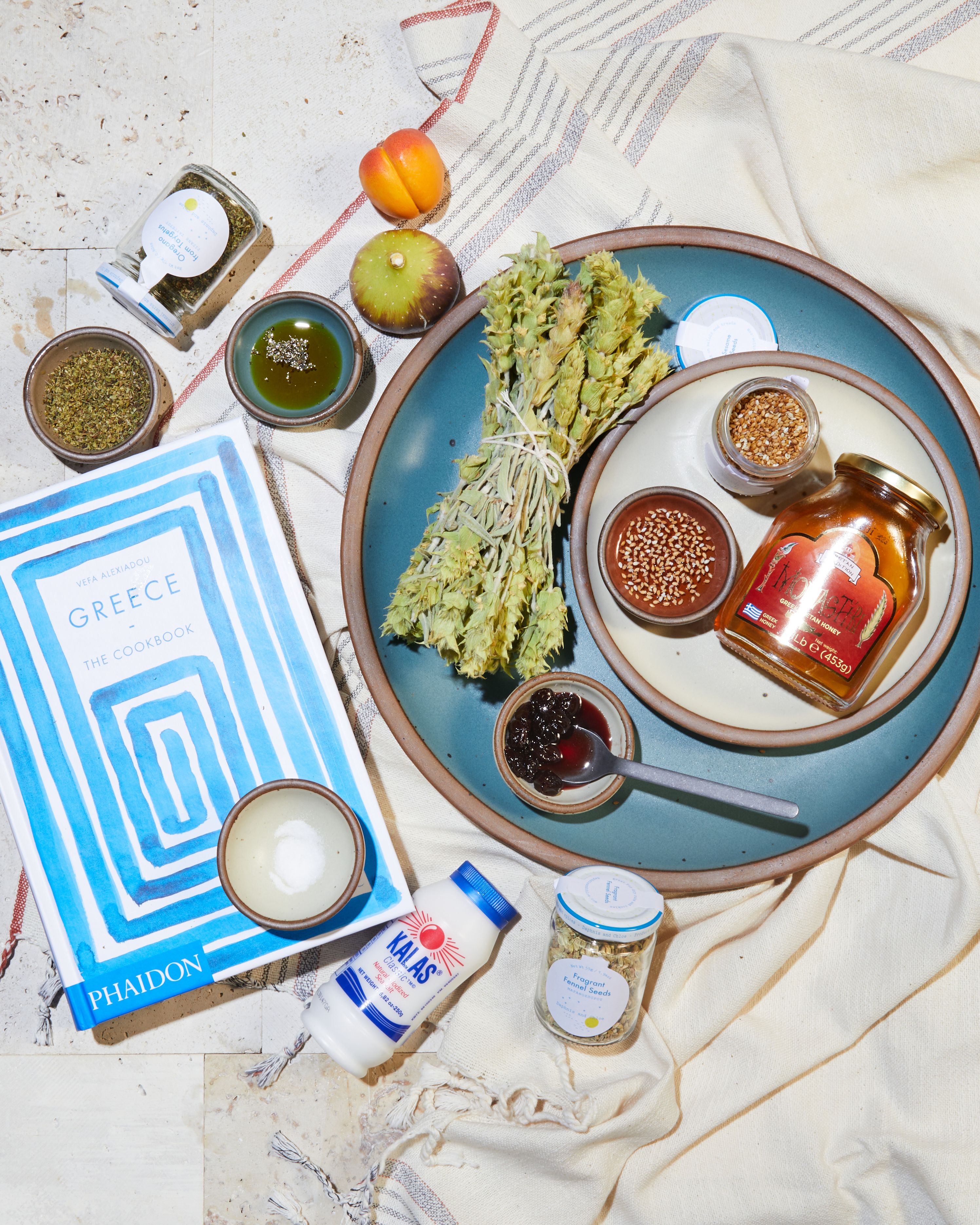 Greek pantry items placed around a Secret Beach dinner plate and Greece: The Cookbook. 