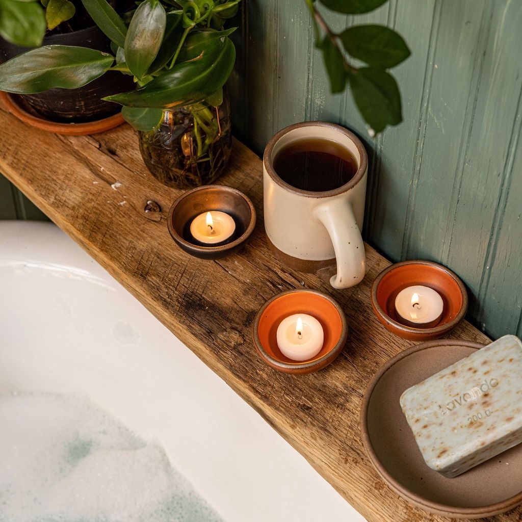 On a shelf by a bathtub, are three tiny ceramic bowls holding tealights in black and terracotta colors, with an East Fork Mug