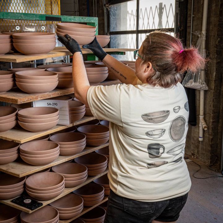 In a warehouse setting, a person is setting a stack of unfinished ceramic bowls on a large shelf. They are wearing a t-shirt with an illustration of ceramic pots on the back.