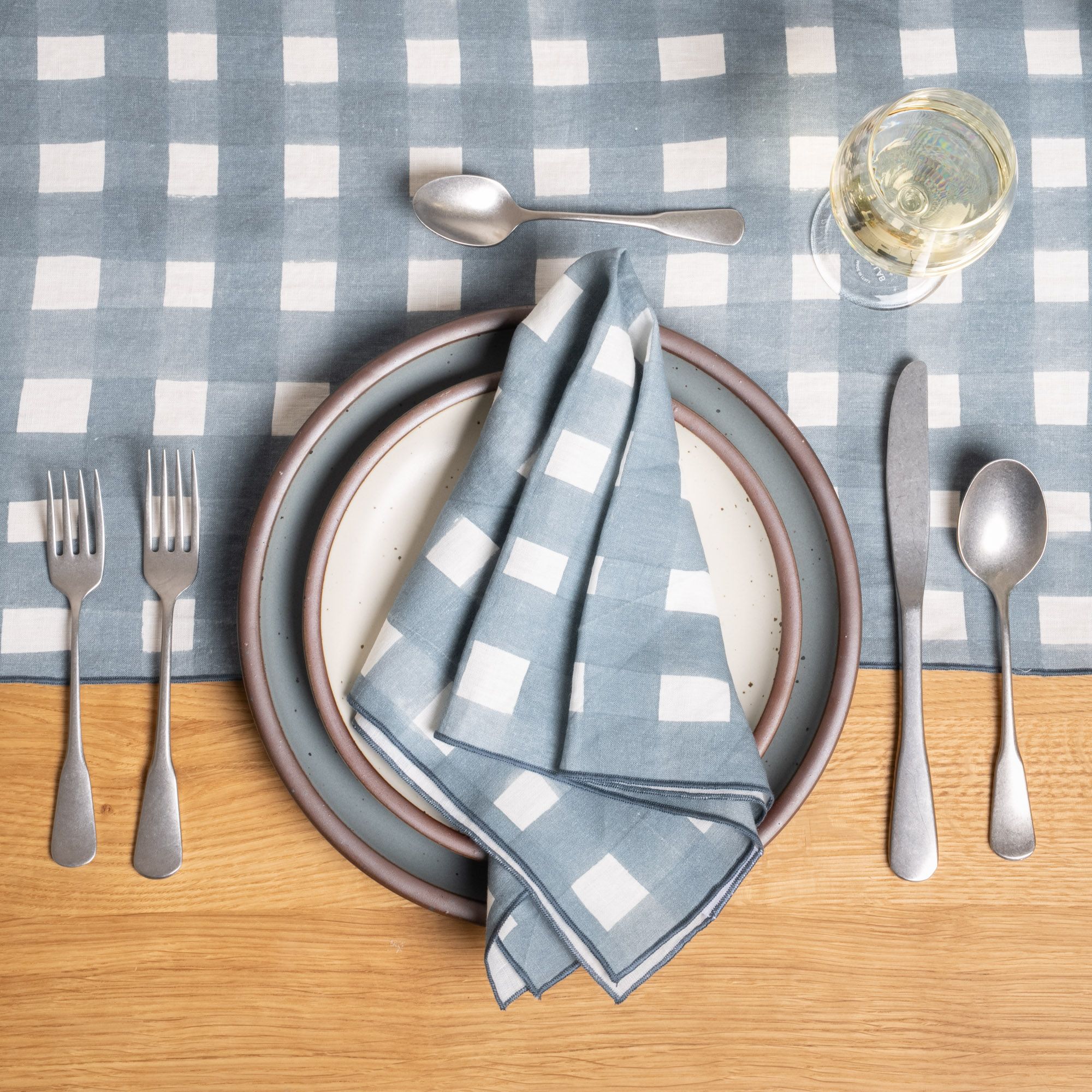 A place setting of a blue-grey and cream ceramic plate, silverware, and wine glass, adorned with a folded blue-grey crosshatch napkin and table runner.