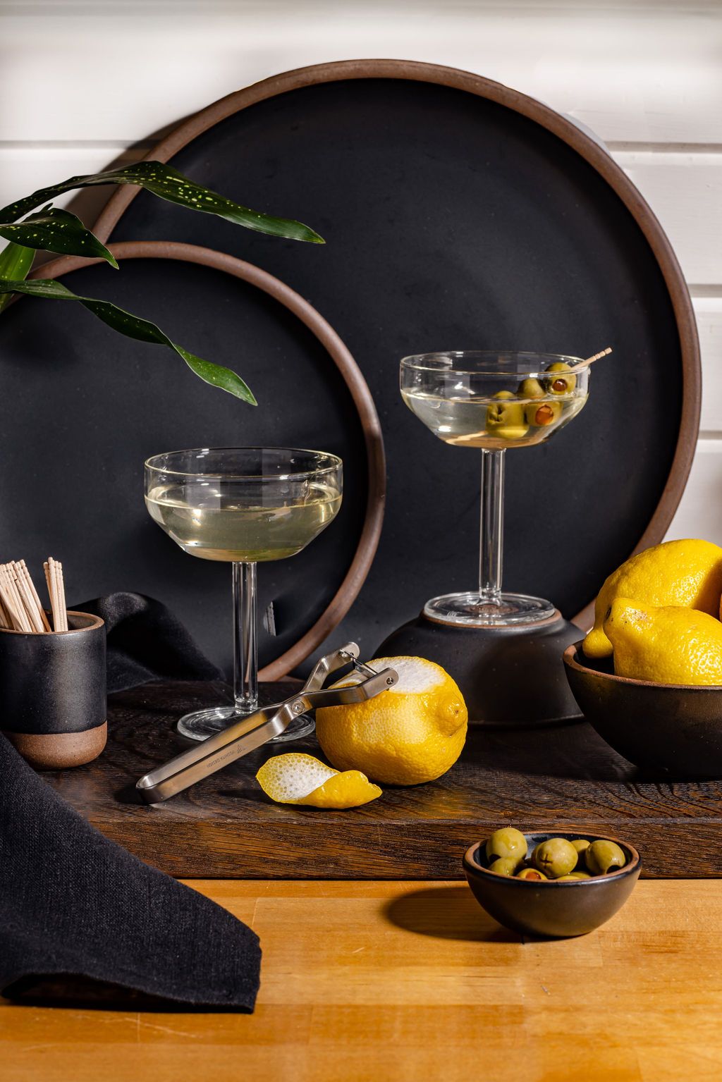 2 full Martini Coupes are artfully arranged with on a counter next to black ceramic pottery, a small bowl of olives, a peeler on a lemon, and a black napkin.