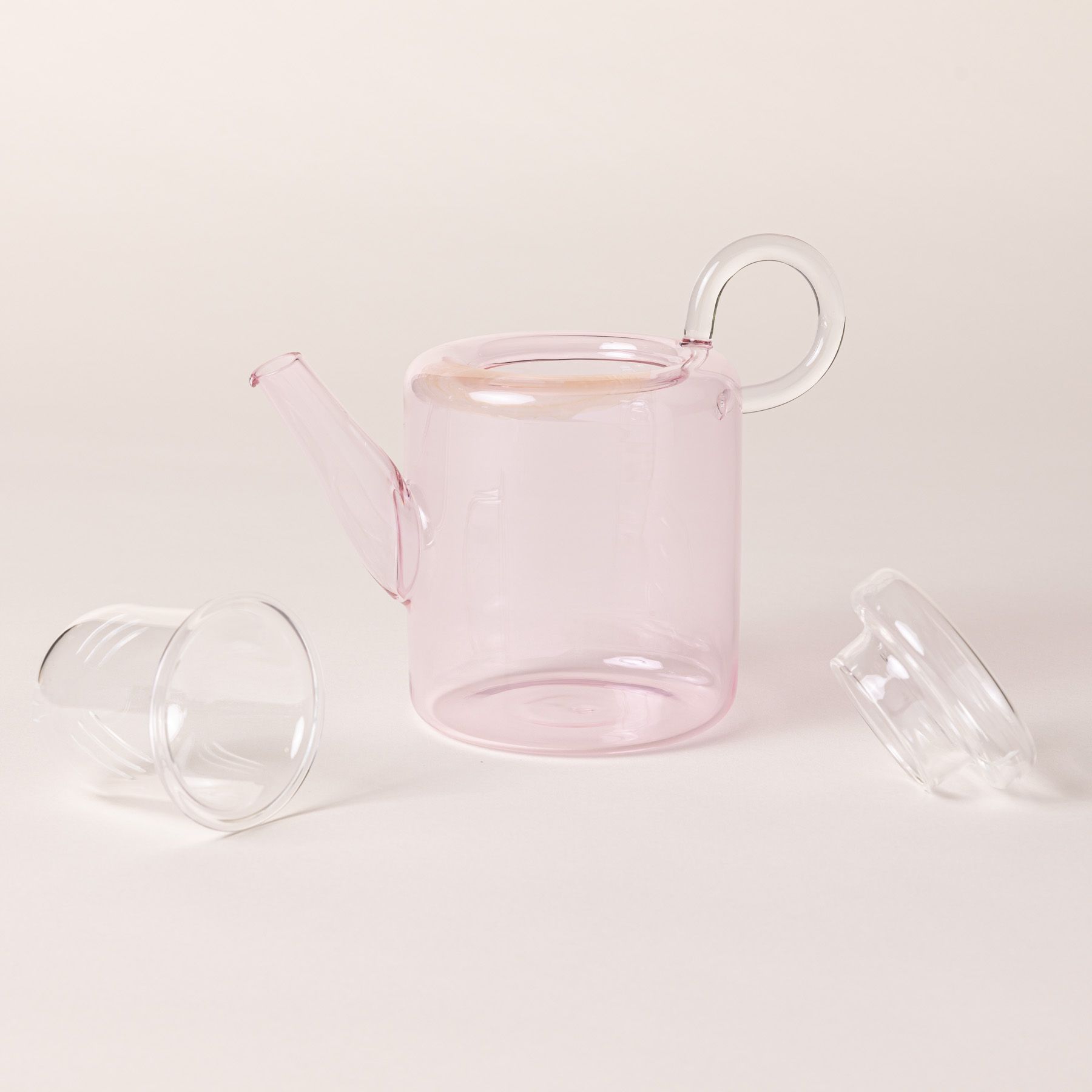 A modern glass teapot in a faint pink color with its strainer and top off to the side.