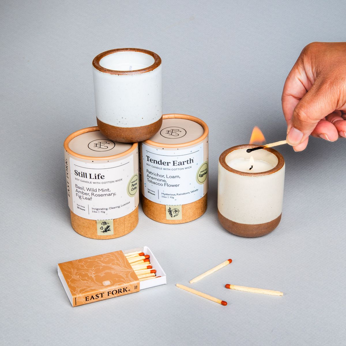 Two small ceramic vessels in cream off-white and cool white colors with candles inside each are gathered next to their cardboard packaging tubes with matches. A hand is lighting the cream candle.