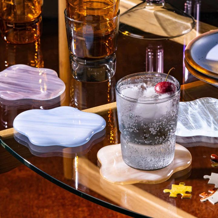 On a table, 4 glass coasters shaped like clouds sit with a simple tumbler filled with a cocktail