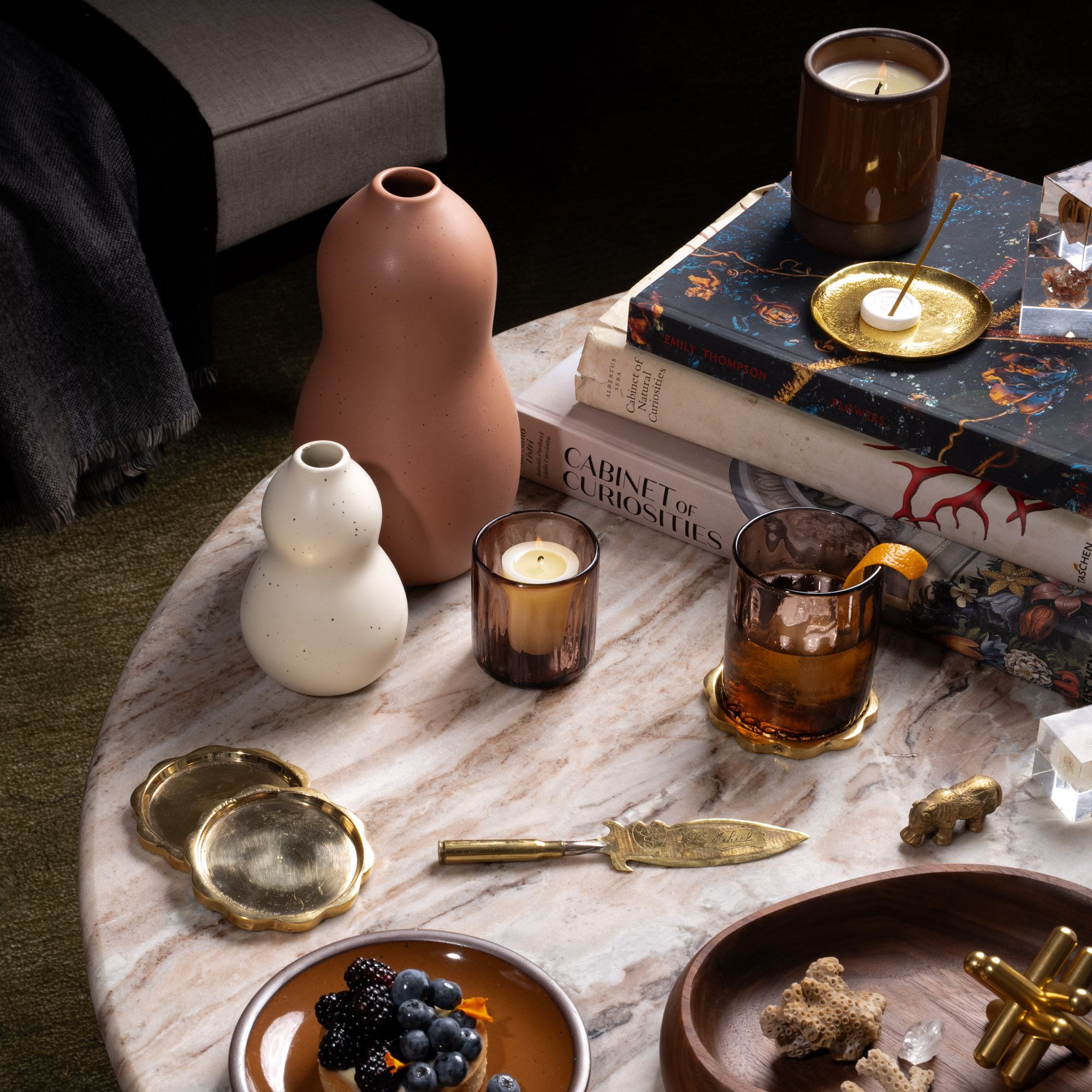 Styled coffee table with sculptural vases, candles, books, brass accents, a drink, and a dessert plate with berries.