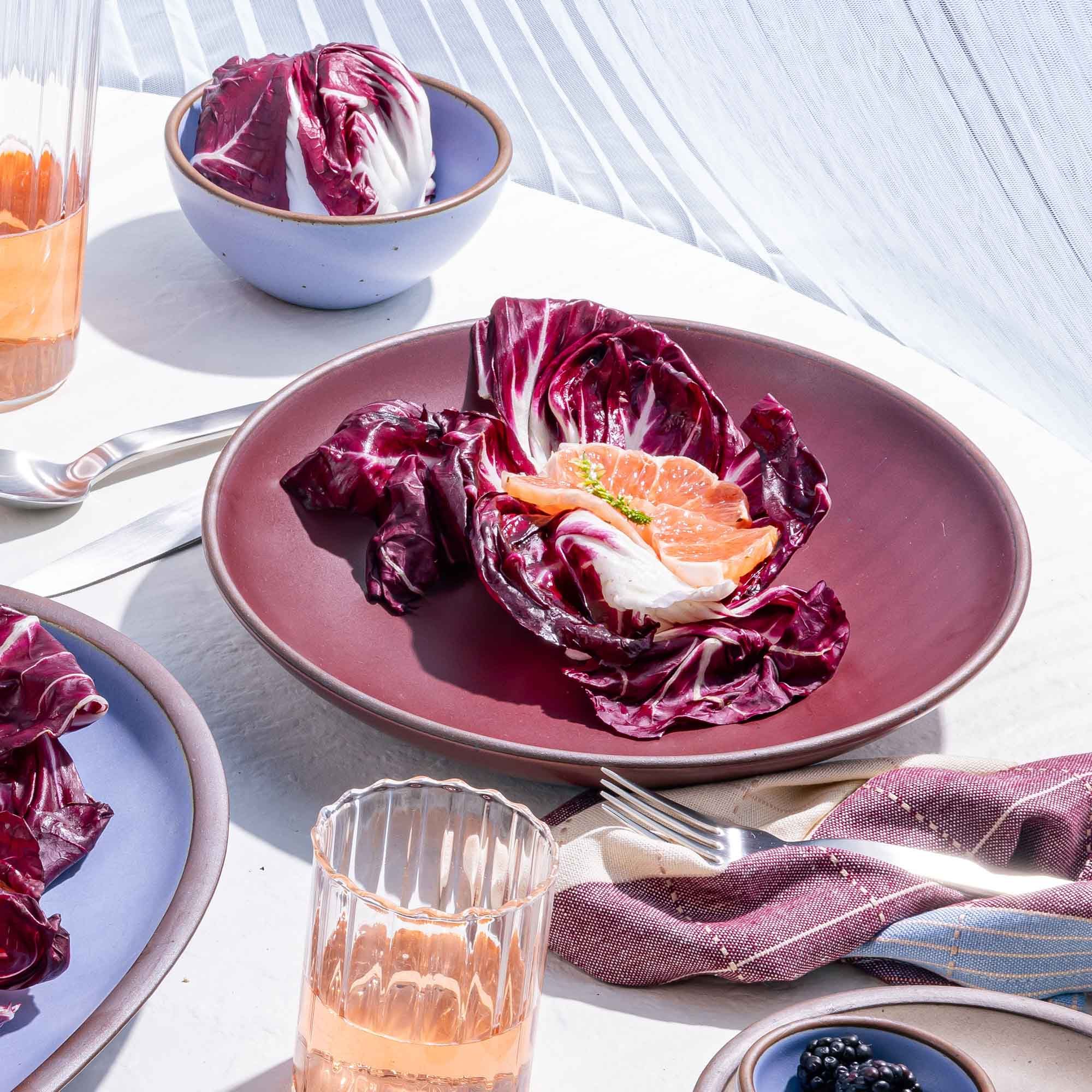 A large ceramic plate with a curved bowl edge in a plum color on a table with red lettuce and glassware