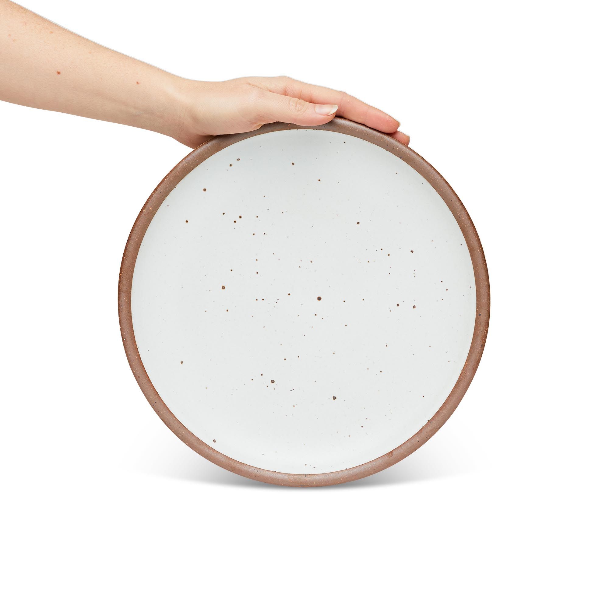 A hand holding a dinner sized ceramic plate in a cool white color featuring iron speckles and an unglazed rim.
