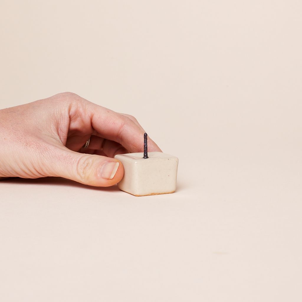 A hand holds a ceramic square candle holder that has a metal spike