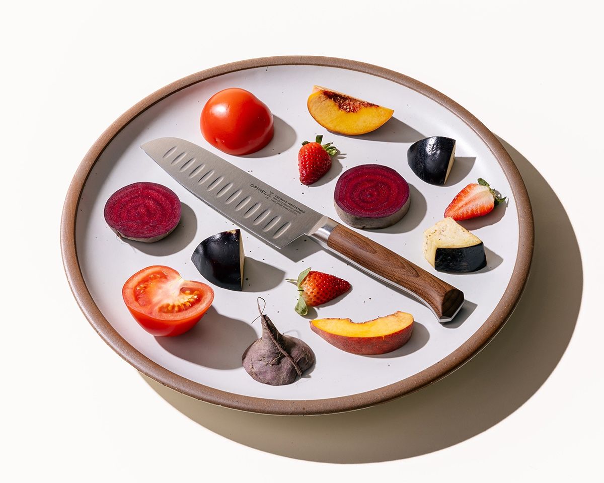 On a white ceramic plate, a santoku knife is artfully arranged in the center surrounded by colorful sliced fruits and vegetables.