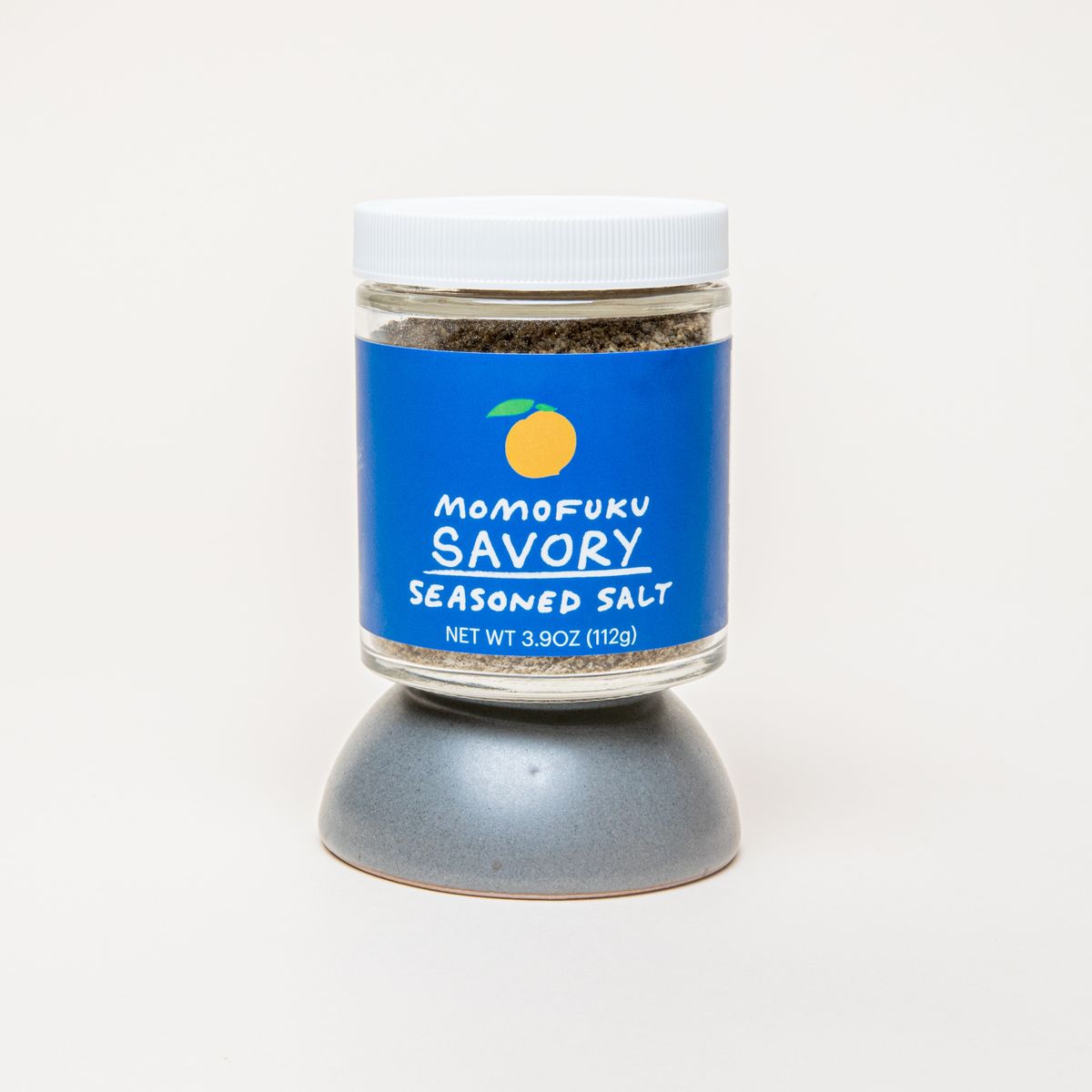 A jar with a white top and blue label that reads "Momofuku Savory Seasoned Salt", sitting on top of an upside down tiny grey bowl