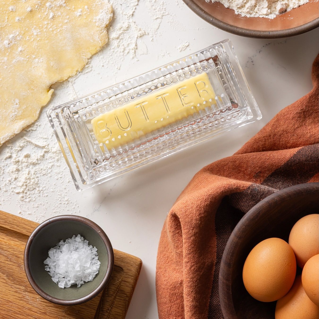 Traditional glass butter dish with 'Butter' on top and butter inside. Off to the side are a bowlful of eggs, a terracotta towel, dough, and a bitty bowl filled with salt.