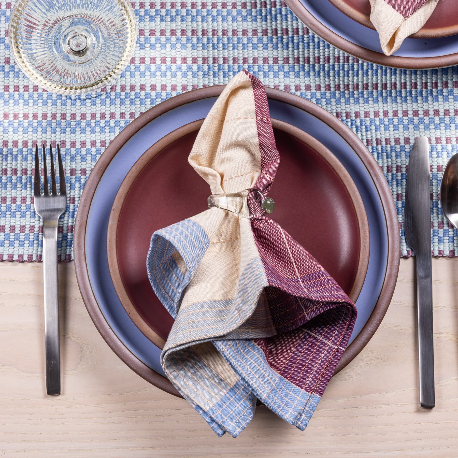 A plaid napkin in periwinkle and plum colors with a glass napkin ring sits on dishes of the same colors with silverware