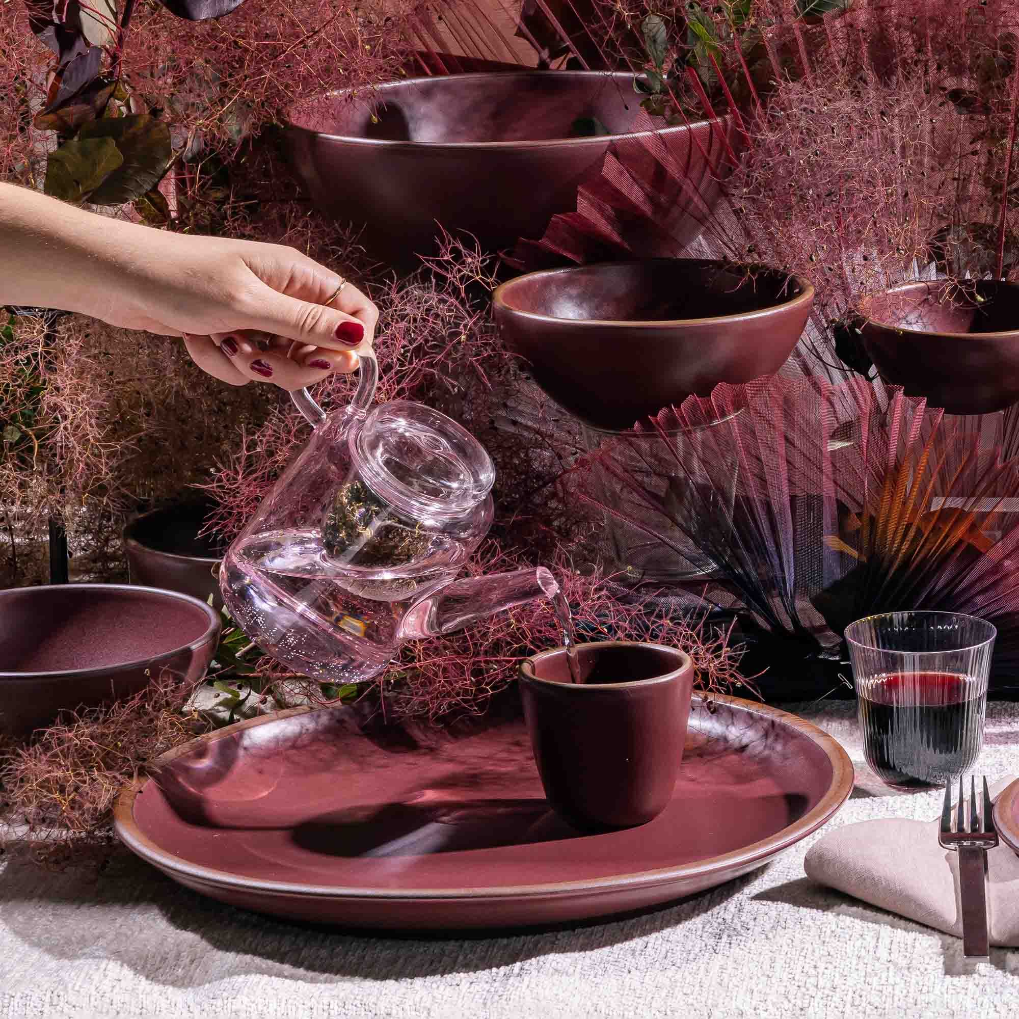 A hand holds a modern glass teacup and pours into a ceramic plum cup surrounding plum pottery.