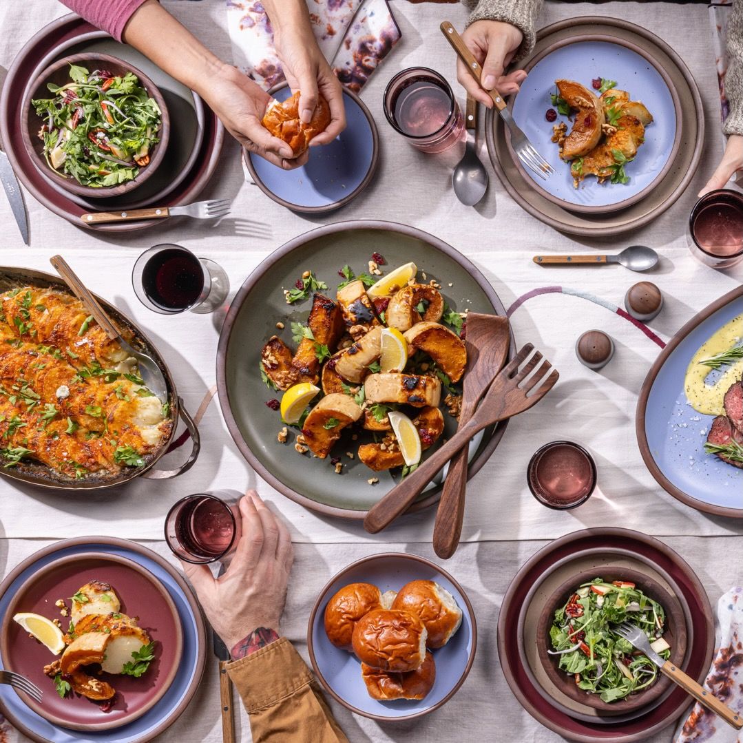 An overhead view of a table filled with a large spread of food on plates and bowls in forest green, periwinkle, and plum colors with wine glasses, flatware, and more.