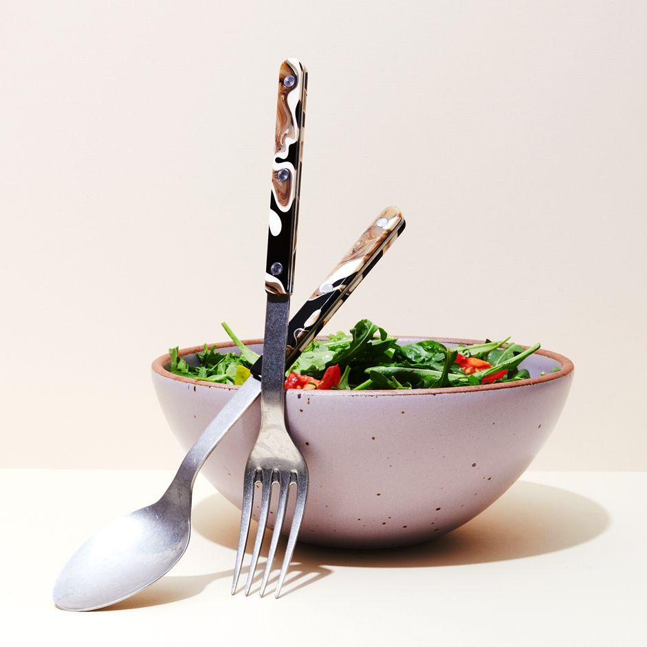 Set of one long spoon and one long fork for serving leaning against a purple bowl containing salad