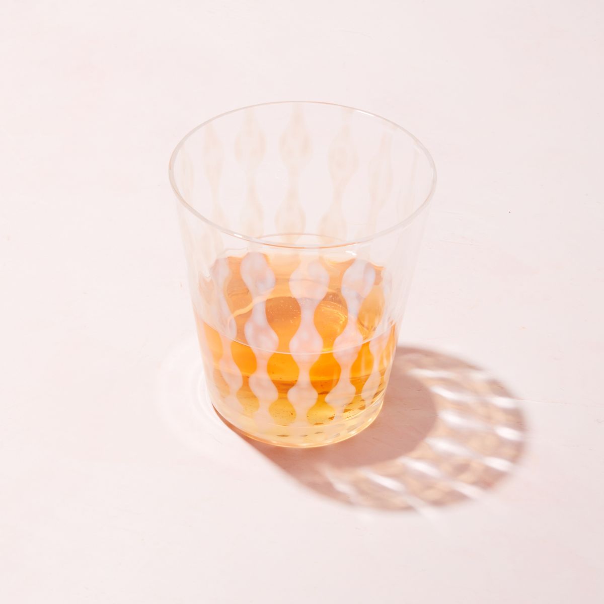 A short clear glass with a delicate vertical wave pattern, half filled with a drink and casting a shadow