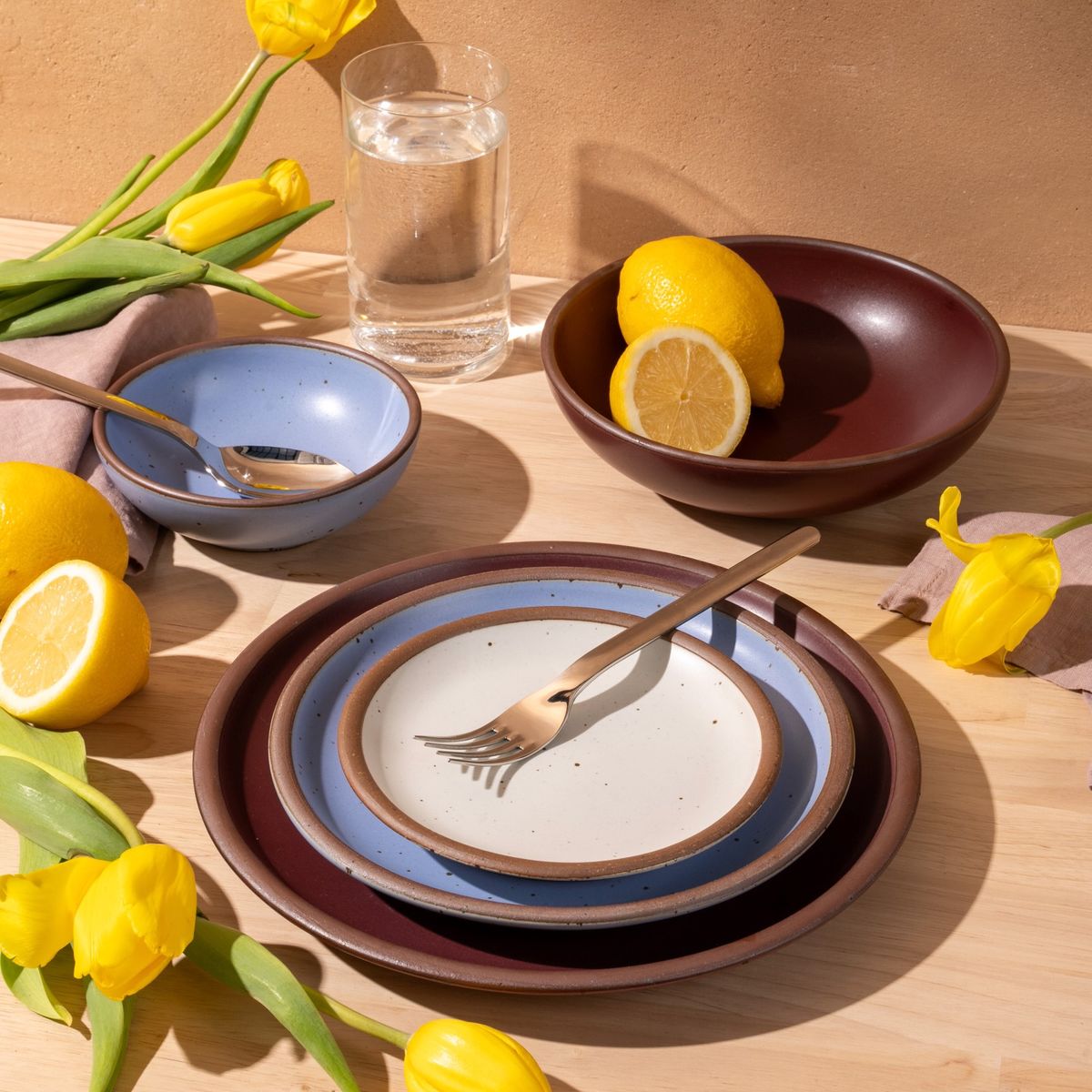 An artful dinnerware set featuring 3 different sized plates and 2 small and large shallow bowls in plum, periwinkle and cool white colors on a table. There is also silverware on the plates, a tall glass of water, and adorned yellow tulips and lemons.