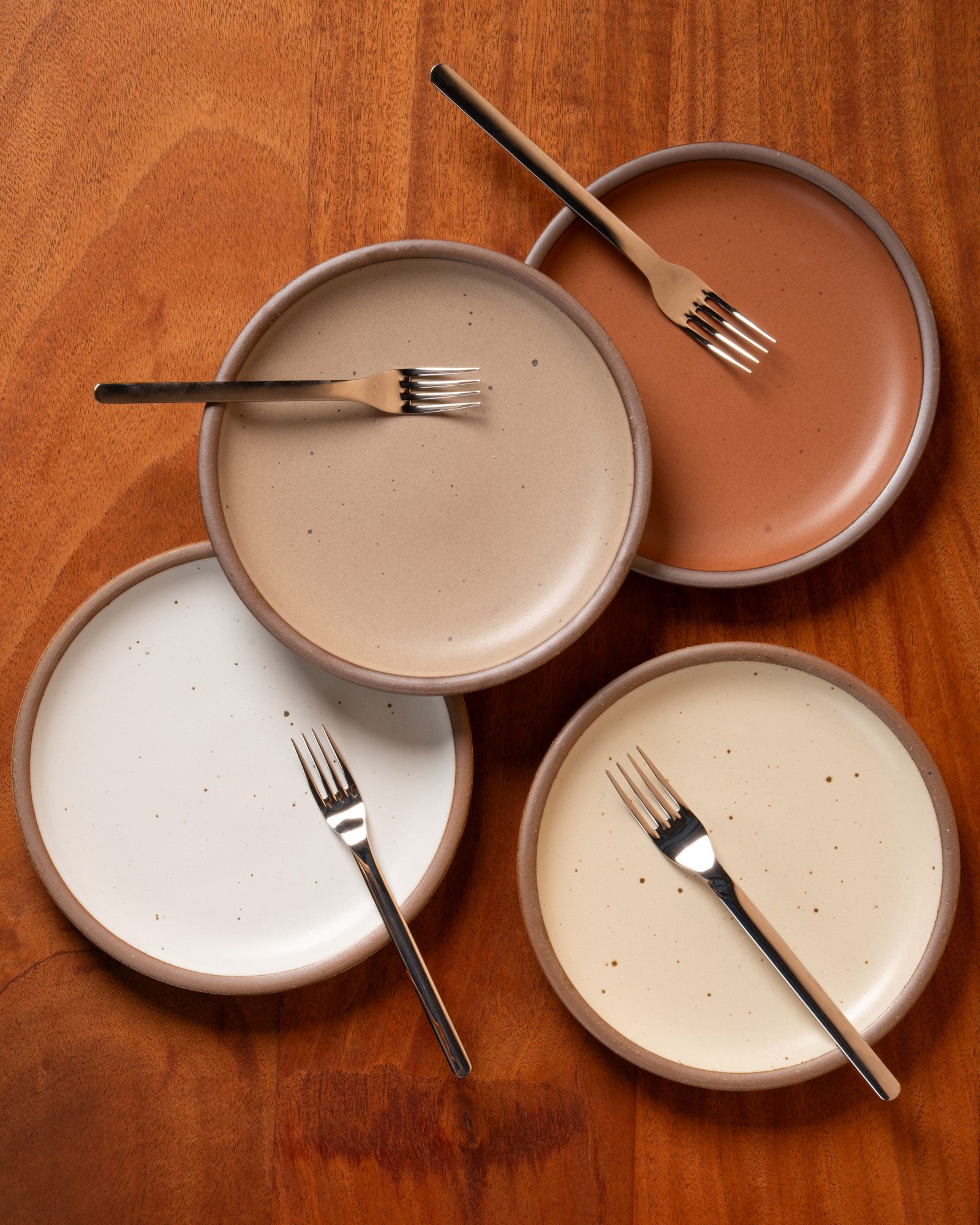 A bird's eye view of 4 ceramic side plates in different neutral colors (white, cream, warm pale brown, and terracotta) each with a silver fork on each plate.