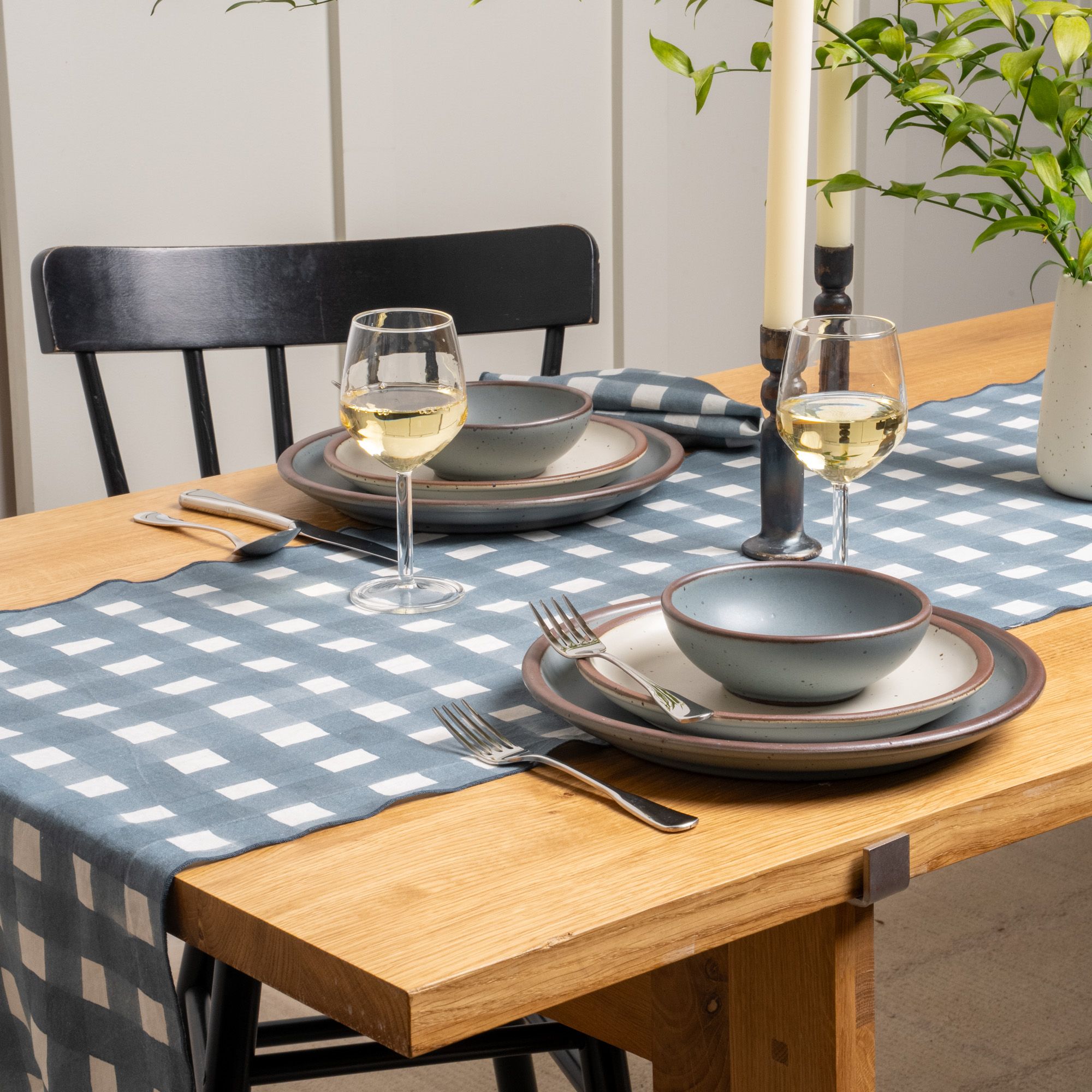 A table featuring a blue-grey crosshatch table runner with 2 place settings of ceramic dinnerware, wine glasses, and silverware.