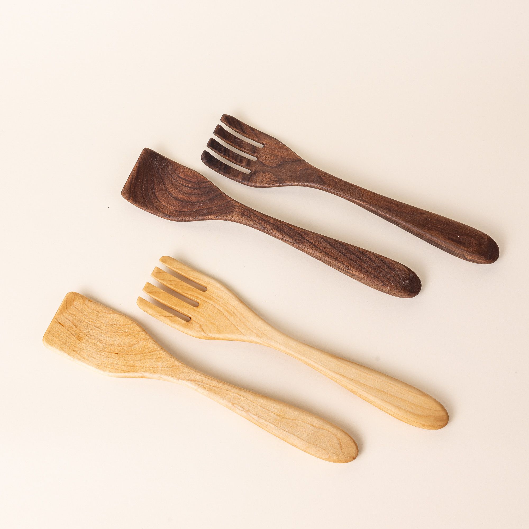 Two pairs of long wood serving utensils in maple and walnut woods. The left utensil has a shape like a spatula, the right has a shape like a fork.