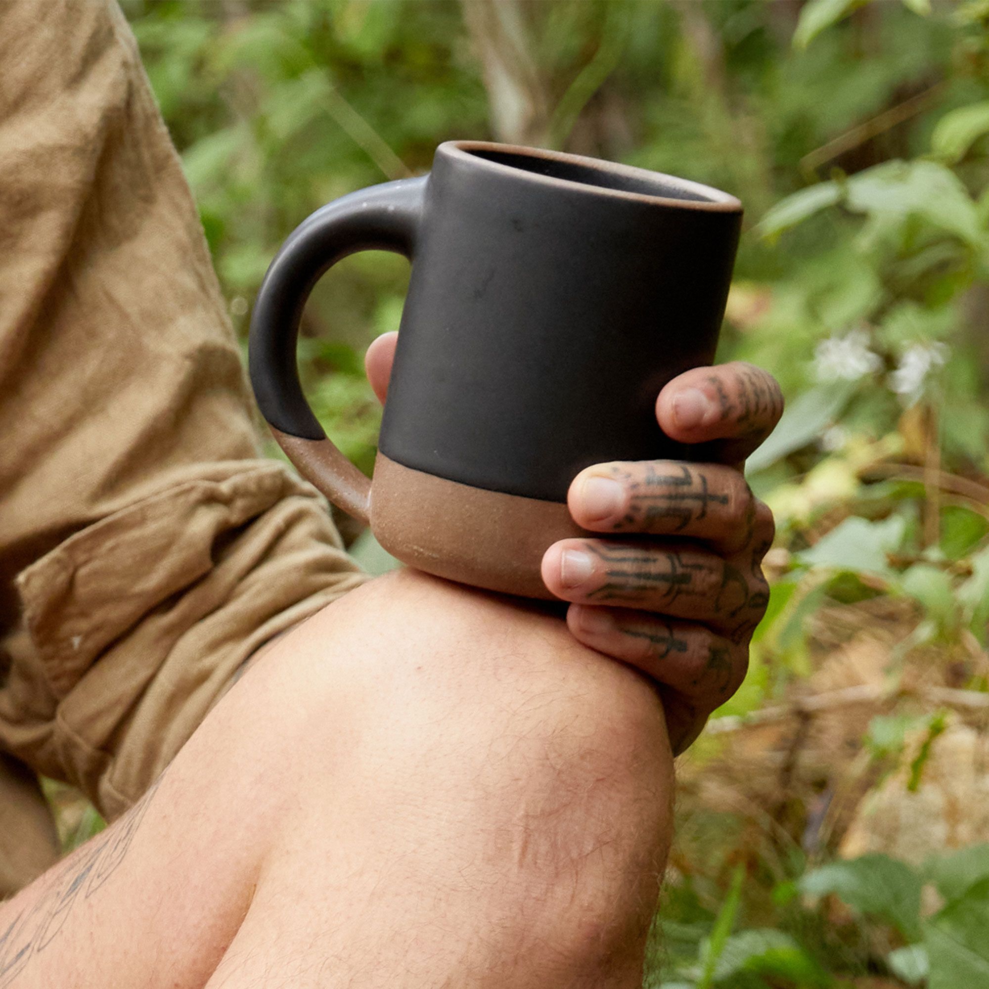 A hand holding a medium sized ceramic mug resting on their knee. The mug is in a graphite black color featuring iron speckles and unglazed rim and bottom base.