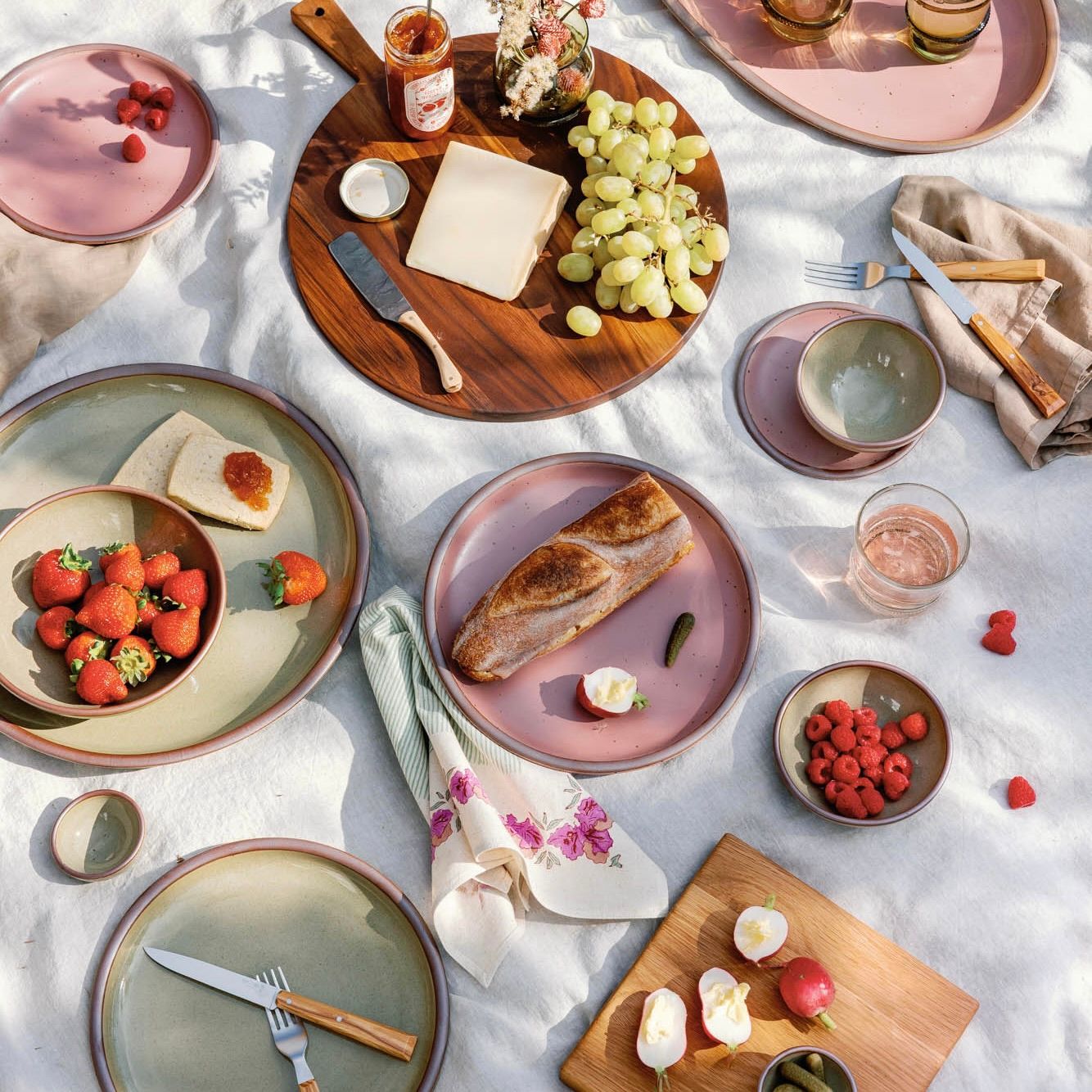 An outdoor picnic filled with ceramic dinnerware in dusty mauve pink and grey-green colors, bread, berries and cheese, a charcuterie spread, glassware and more.