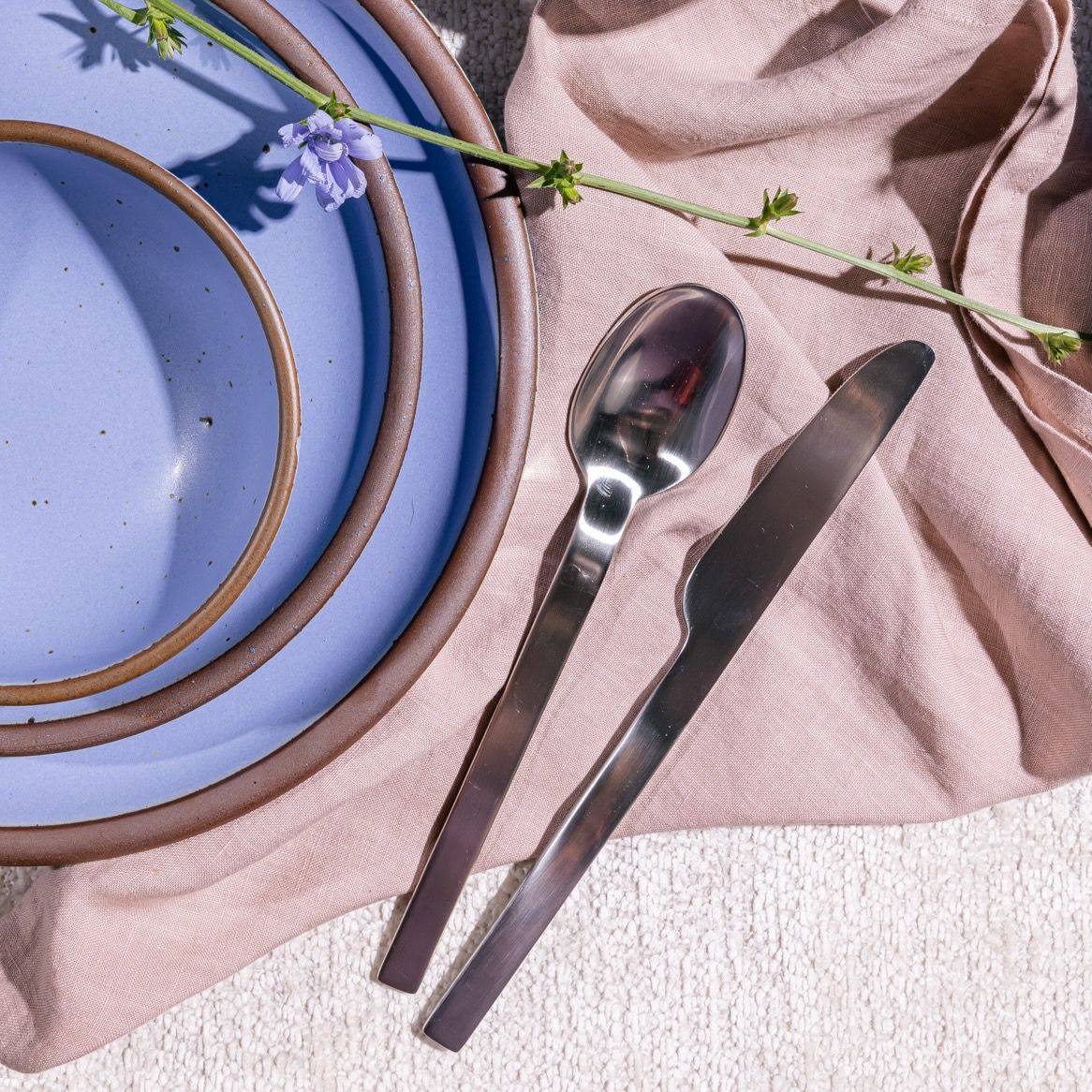 A simple steel spoon and knife on a light pink napkin next to ceramic dinnerware in a periwinkle color
