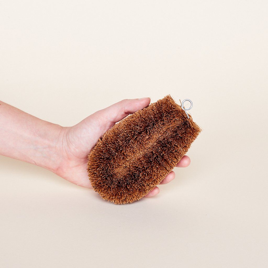 A palm hemp fiber oblong scrub brush with medium and dark brown bristles held by a hand