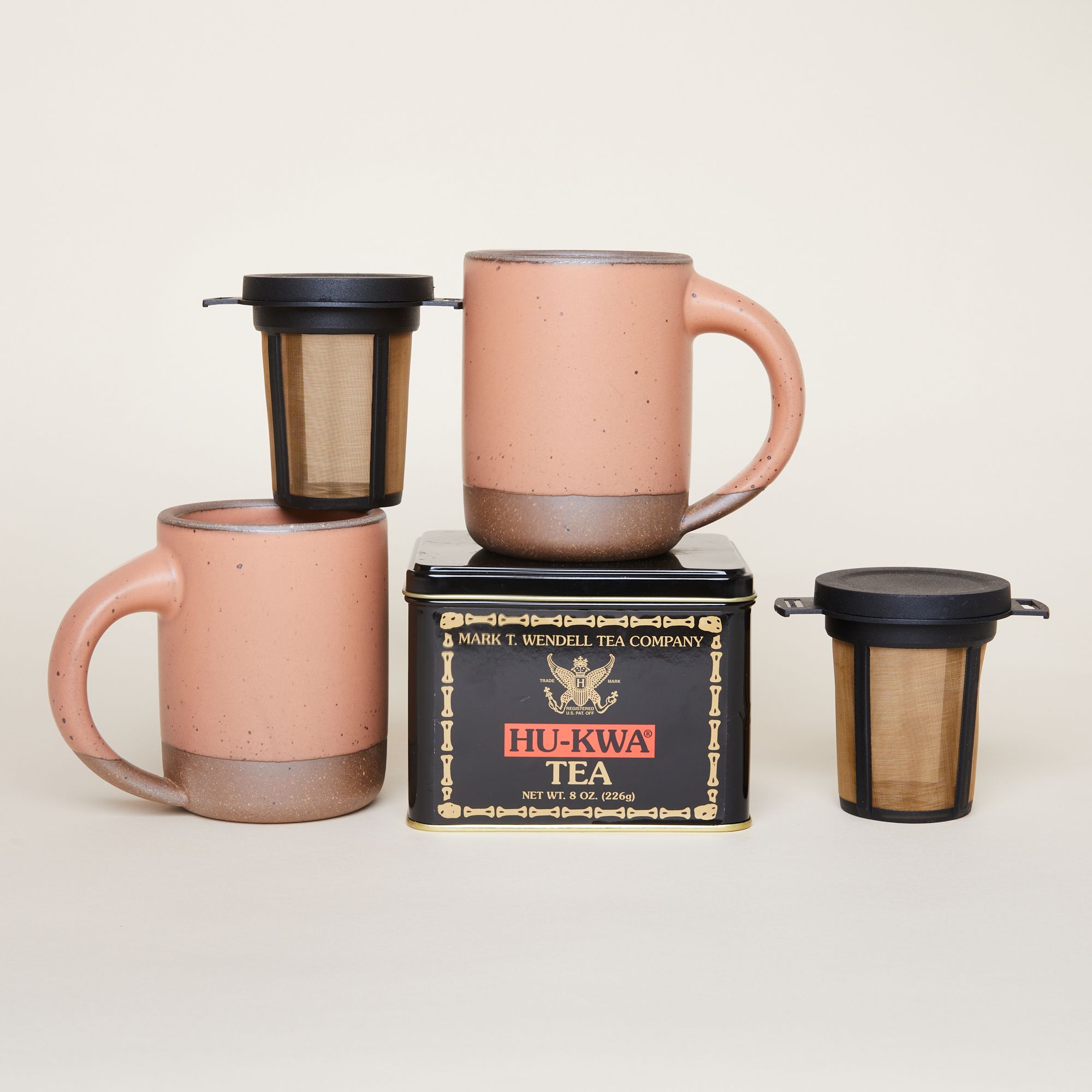 Two East Fork Mugs in Utah, a warm, soft, pinkish orange, sitting next two a black and mesh tea strainer that fits right in a mug and a black box of black tea.