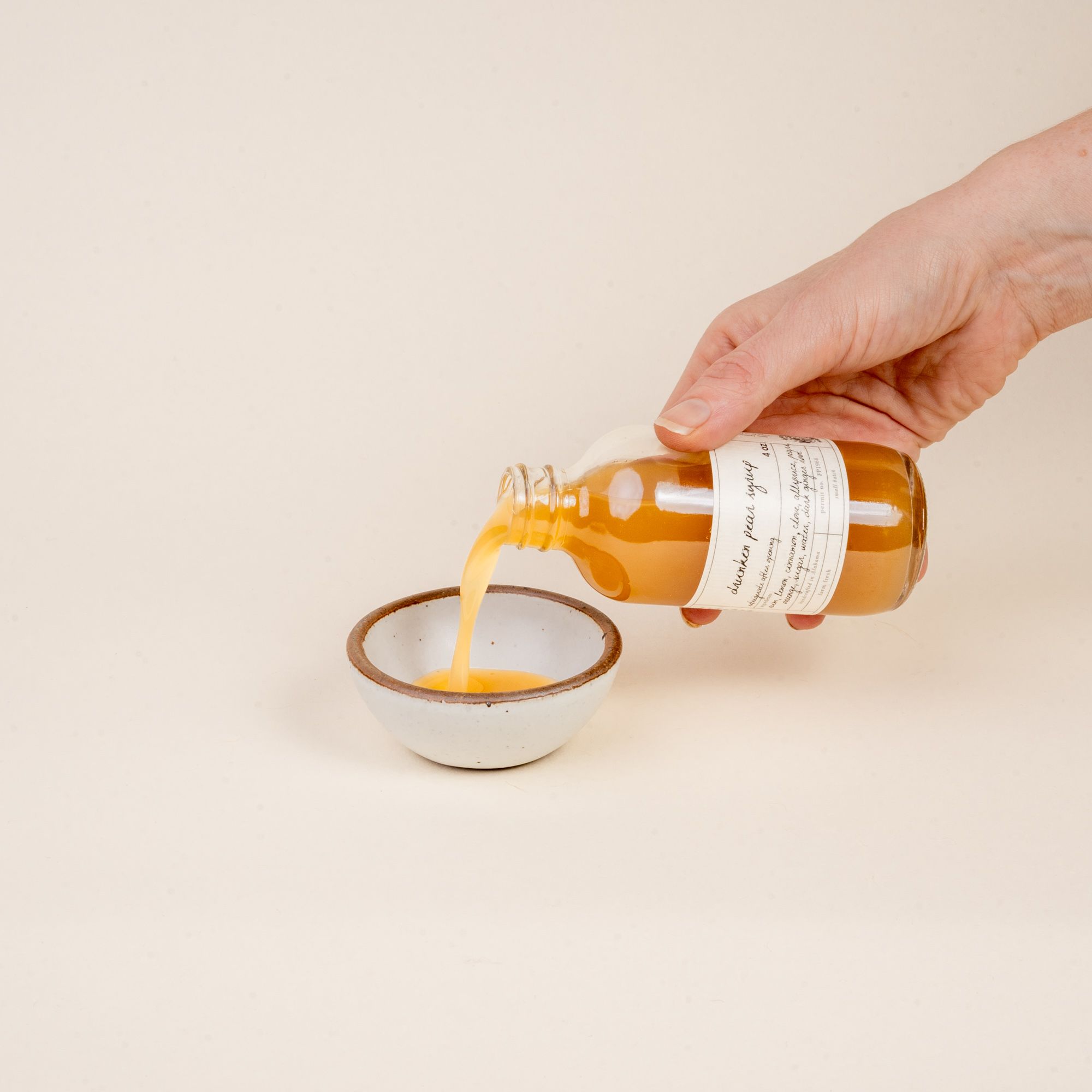A hand pours out some syrup into a small ceramic bowl out a small bottle that reads 'Drunken Pear Syrup'