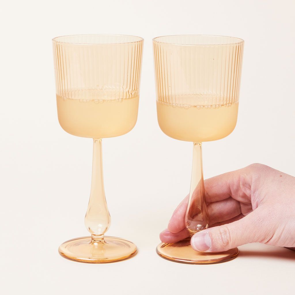 A hand rests on the base of a clear yellow wine glass that is filled parting with a clear liquid. An identical glass stands to the left