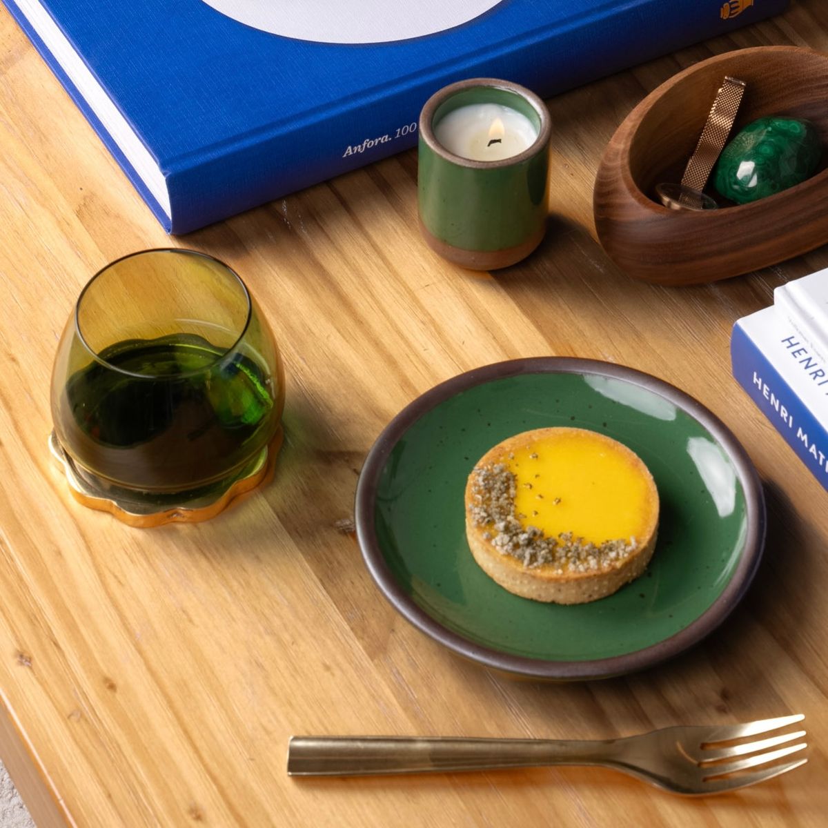 Glossy green plate with a yellow tart, green whiskey glass, candle, books, and wooden dish on a wooden table.