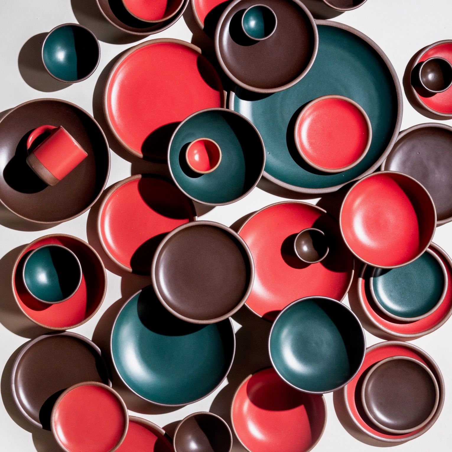 A bird's eye view of ceramic bowls and plates in bold red, dark teal, and dark brown colors. They are in all different sizes stacked and arranged artfully.