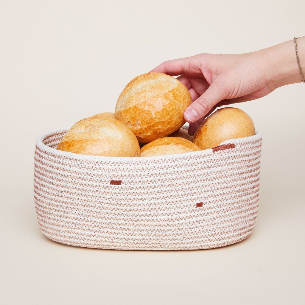 Hand reaching for bread roll among many bread rolls resting in a woven bread basket with red trim.