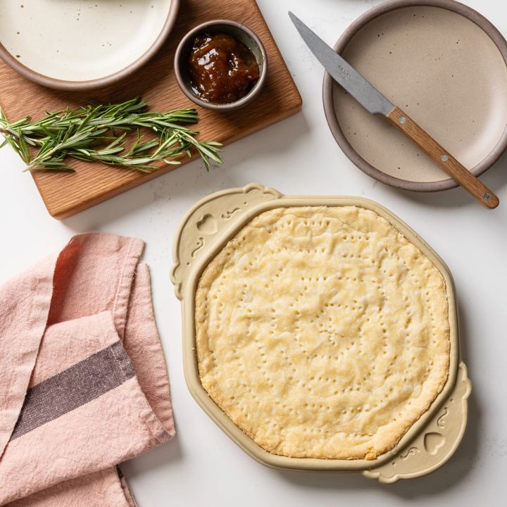 On a countertop, there's a baked shortbread in a shortbread pan, a plate and knife, a napkin, along with rosemary and jam.