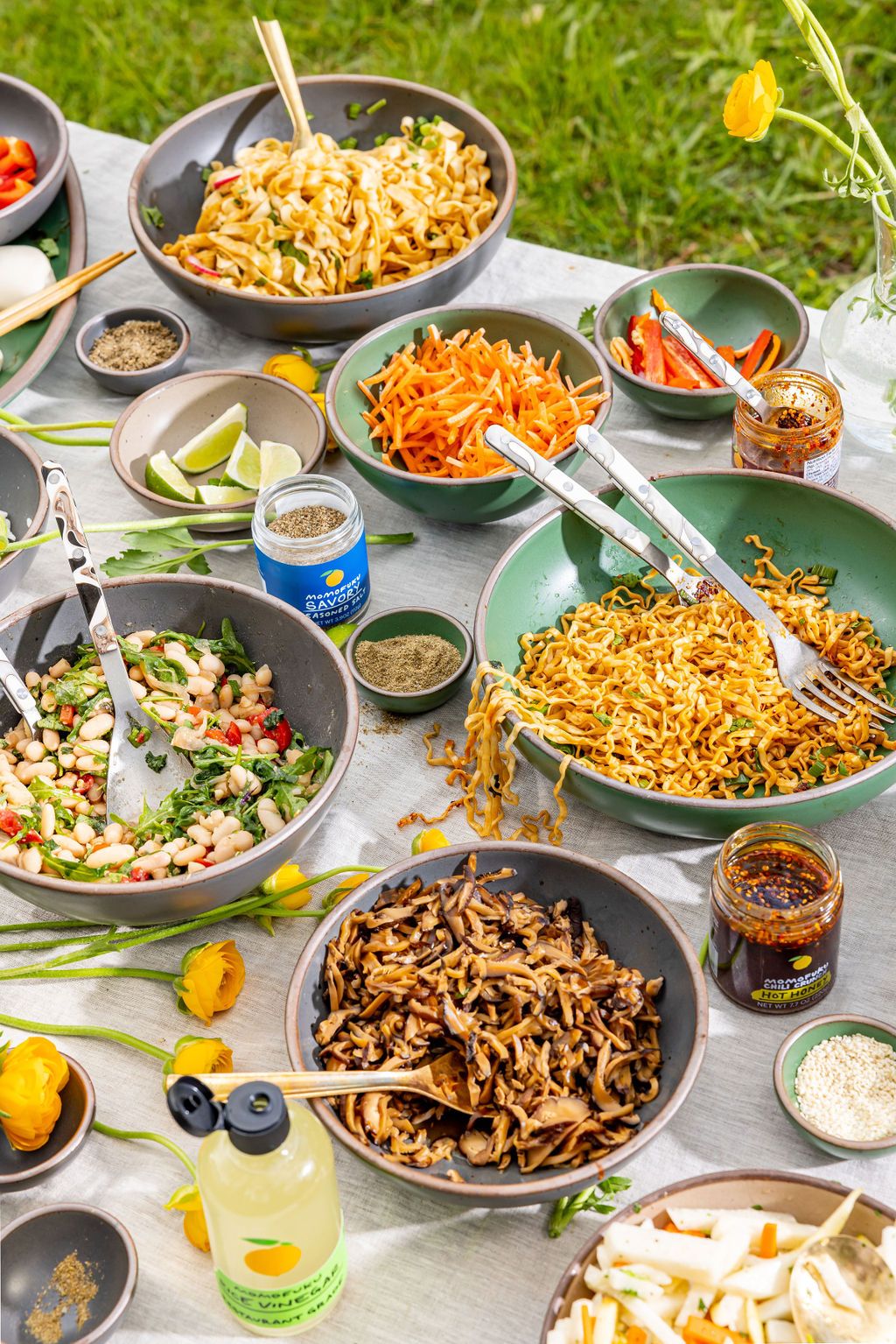 A table takes up most of the frame featuring many grey and green bowls of varying sizes with foods and serving utensils. Scattered around the table between the bowls are a bottle of rice vinegar, small jars of chili crunches, and yellow flowers. There is grass in the background around the table.
