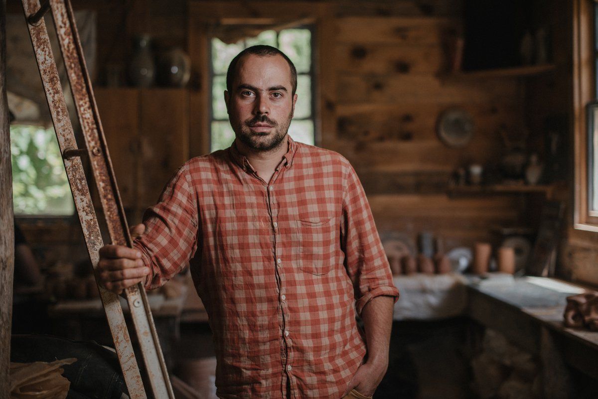 Alex wears a flannel shirt and is slightly leaning against a ladder in a cabin