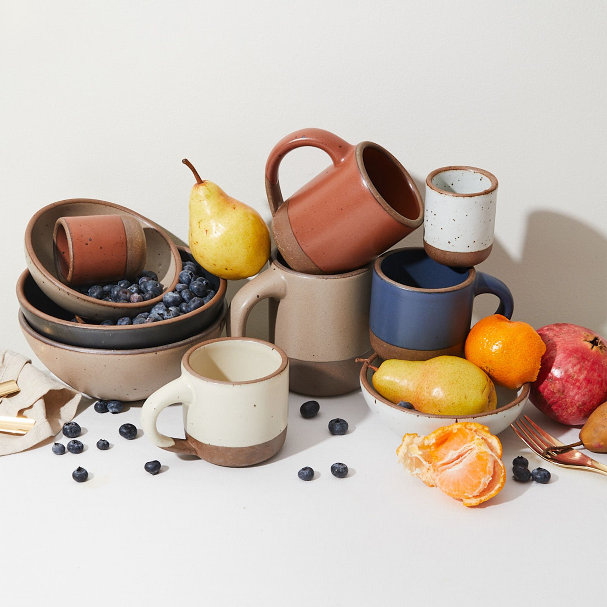 A styled photo of stacked bowls and various mugs and cups along with fruits, napkin, and fork