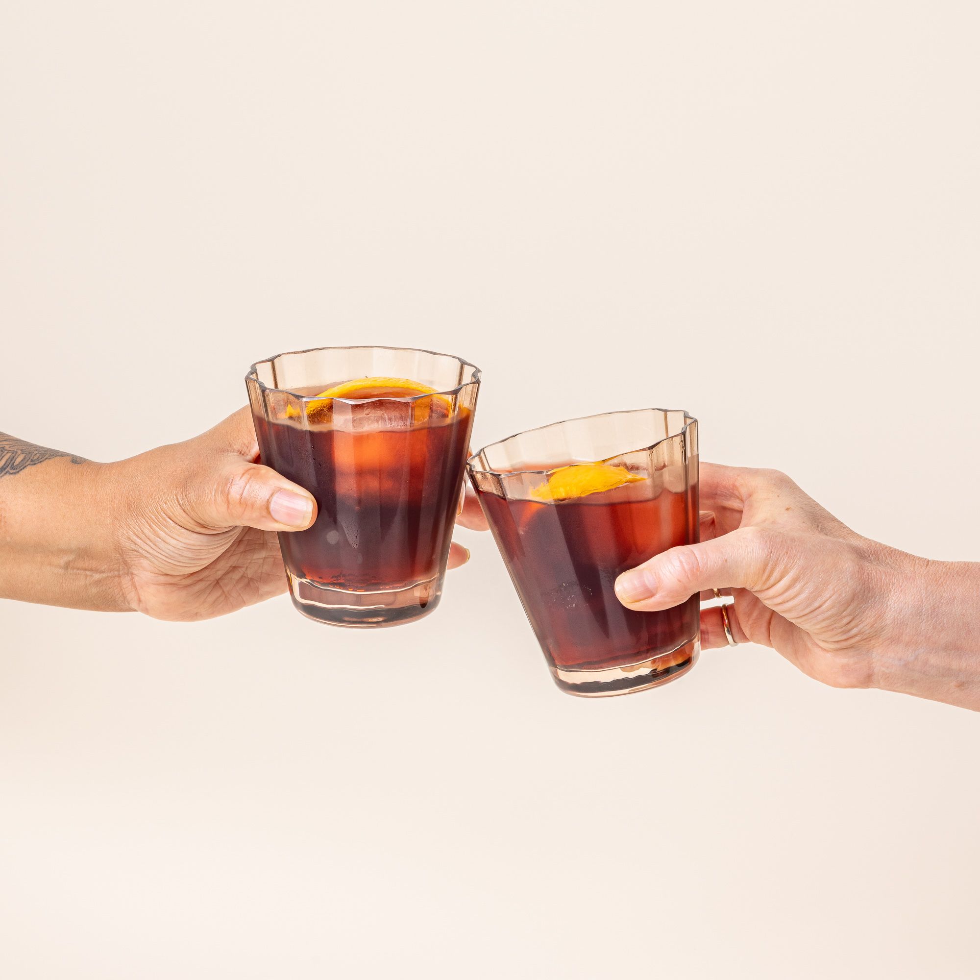 Two hands cheering and holding two transparent light amber glasses with wide grooves on the side, filled with a cocktail and orange slice.
