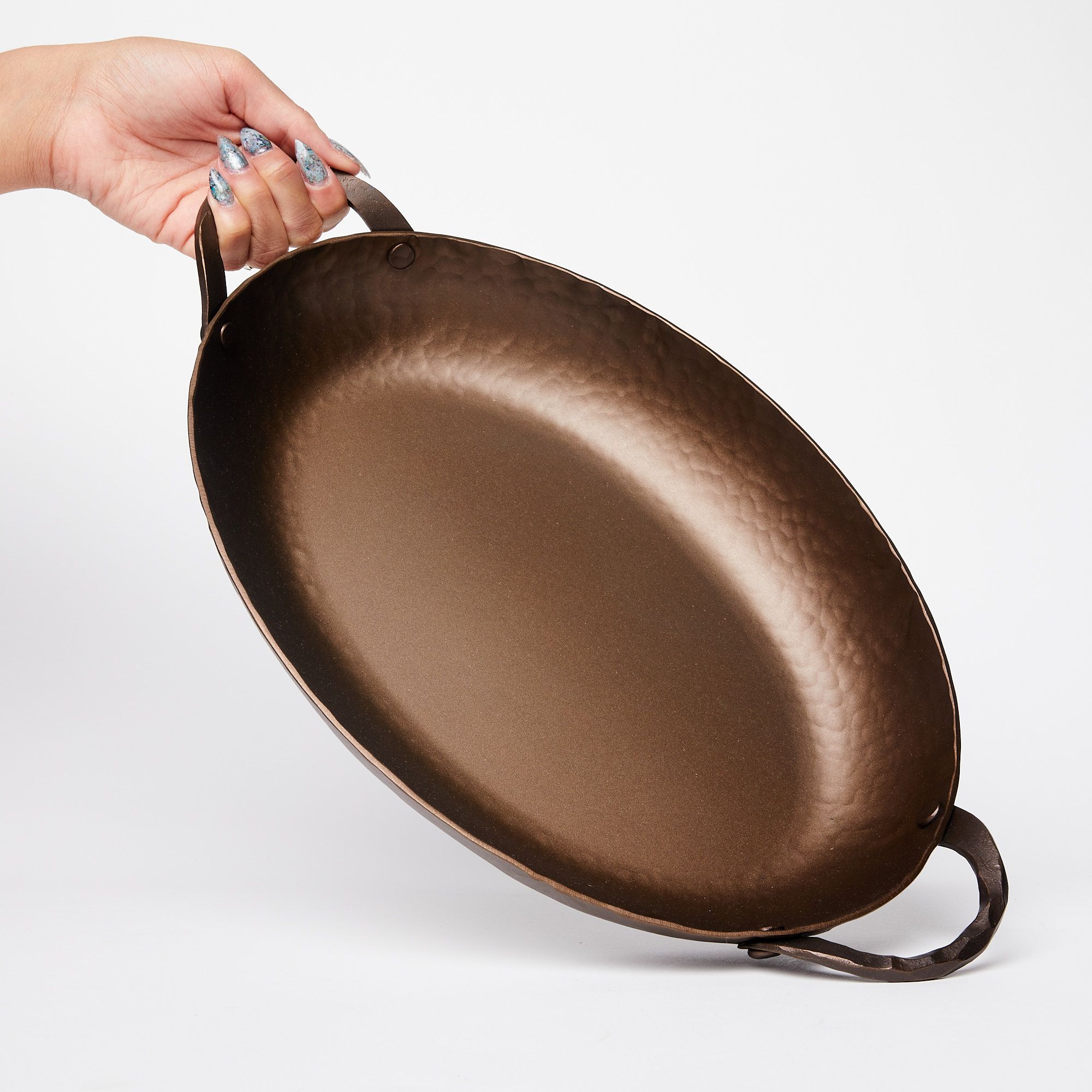 A hand tilting a hammered metal oval roasting pan with handles on both sides