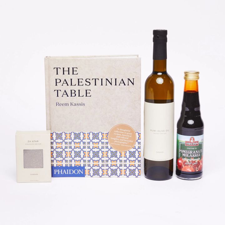 From left to right: A cream-colored box of a za'atar, a hardcover book that reads "The Palestinian Table", a tall brown bottle with a cream-colored label that reads "Rumi Olive Oil", a medium clear bottle full of dark pomegranate molasses 