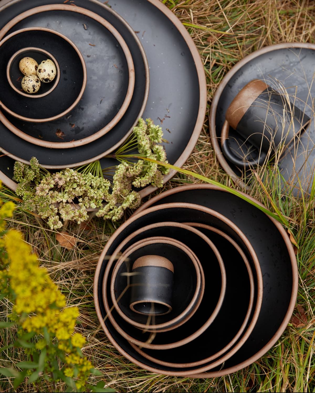 Outside sitting in the grass, is a artful stacks of ceramic plates and bowls in graphite color with unglazed rims.