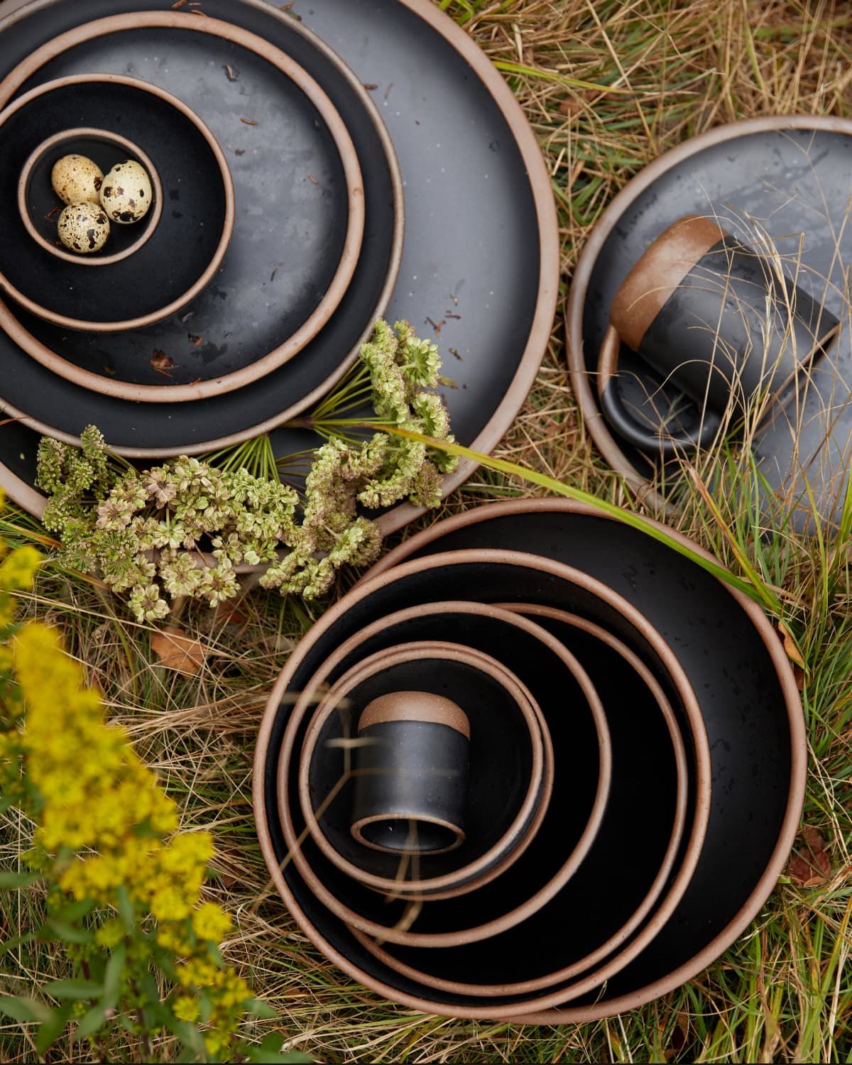 Outside sitting in the grass, is a artful stacks of ceramic plates and bowls in graphite color with unglazed rims.
