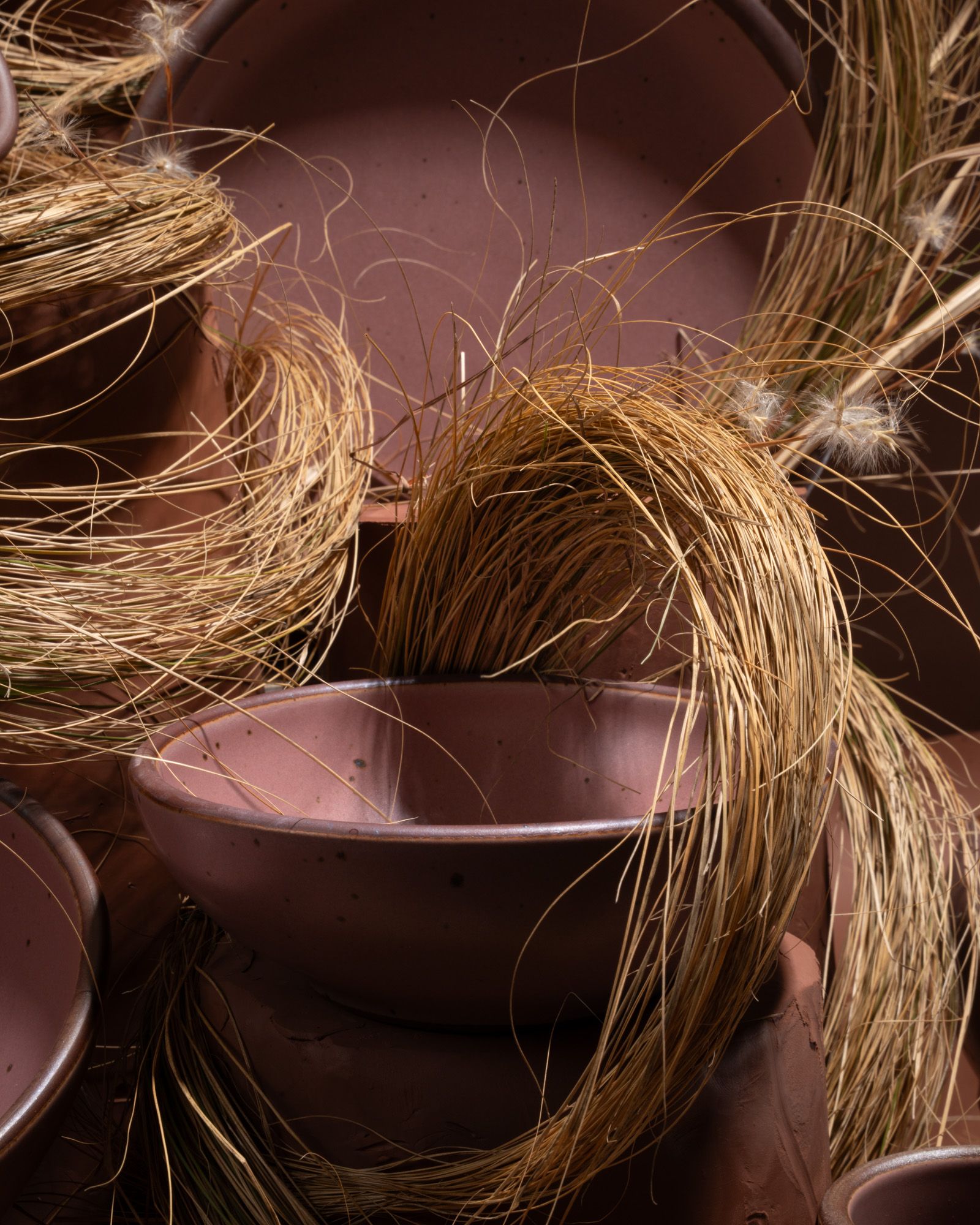 Closeup of a stack of pink ceramic bowls and plates piled together with strands of dried grass against a dark background.
