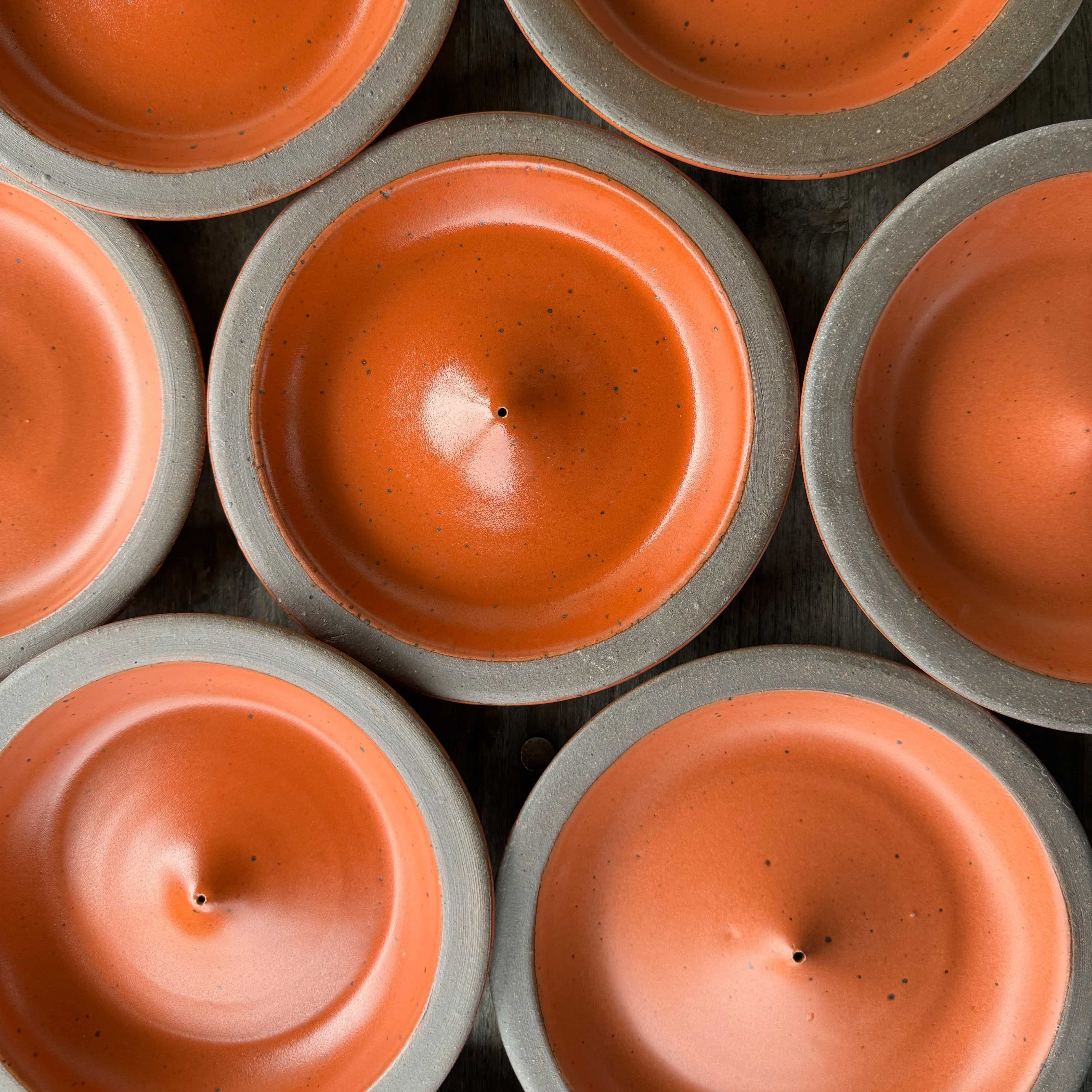 A batch of ceramic plate-shaped incense stick holders with an unglazed rim and glazed in a bold orange color featuring lots of iron speckles.