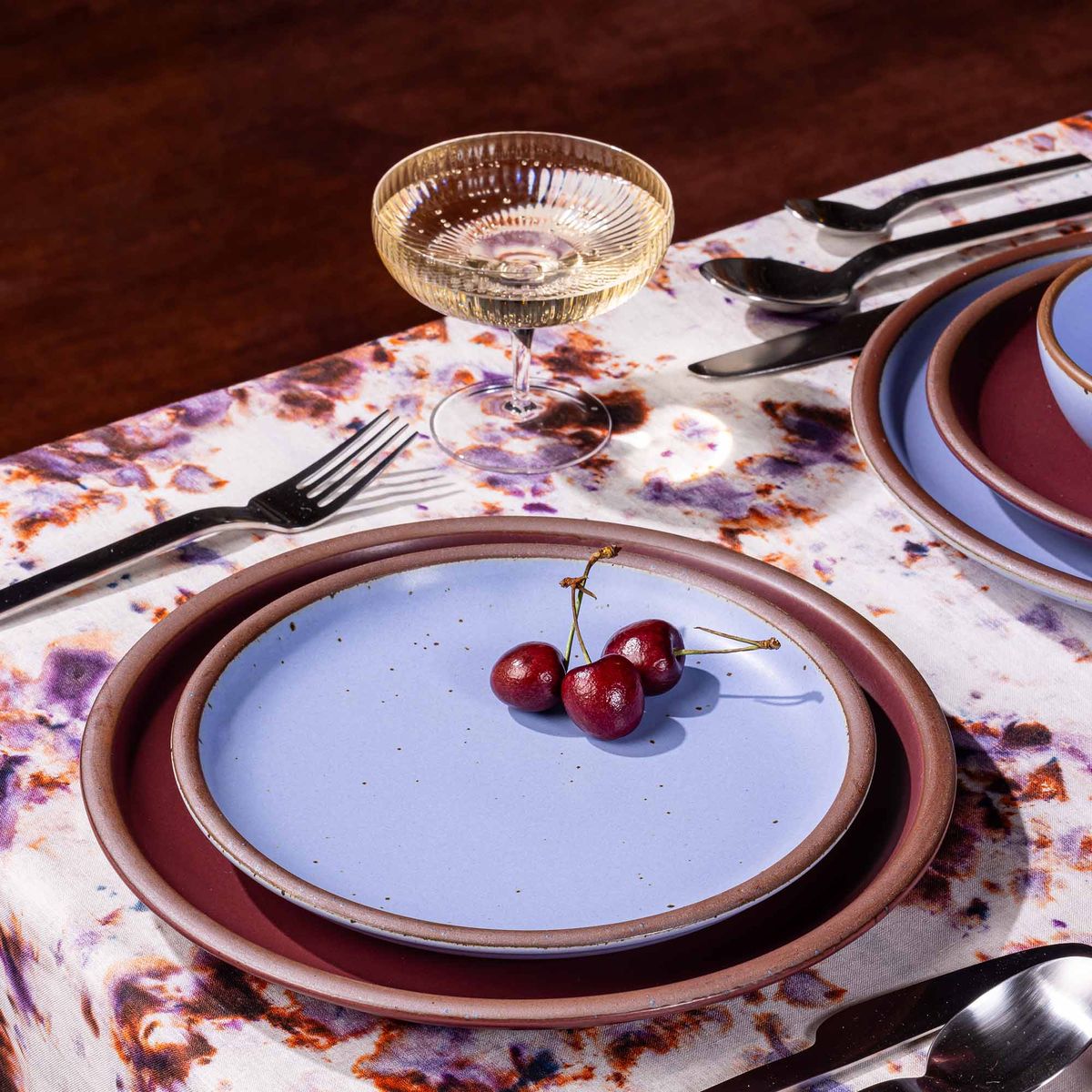 A table setting featuring marbled textiles, a coupe glass, and plates in plum and periwinkle colors