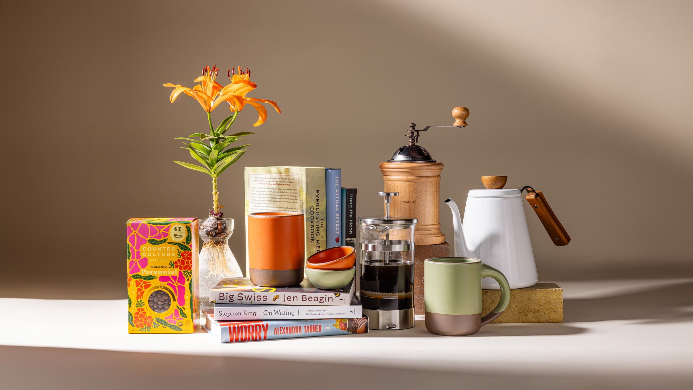 A lineup of various goods all together including ceramic mugs, a clear bulb vase, a stack of books, some coffee accessories like a french press, and a box of coffee.